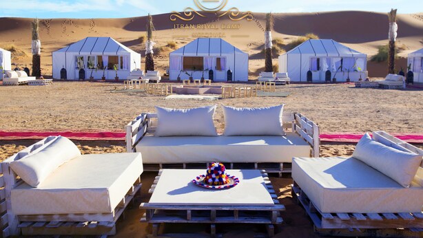 A desert camp with white tents, a lounge area with white sofas and a colorful hat, and a clear view of sand dunes in the background. - Itran Royal Camp (Taouz)