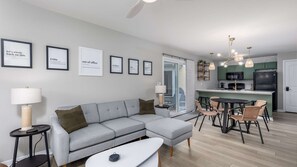 A modern living room with a grey sectional sofa, a dining area with wicker chairs, a kitchen with green cabinets, and framed motivational quotes on the wall.