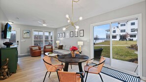 A modern living room with a round table, orange chairs, a sofa, and a large window offering a view of a white building and a grassy area.