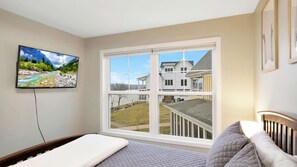 A bedroom with a large window, a bed, a TV, and a view of a house.