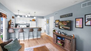 Una cocina moderna con armarios blancos, un microondas y una estufa. Hay tres taburetes de bar y un televisor montado en la pared.