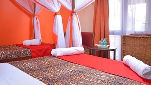 Habitación de hotel con dos camas, una colcha de cuadros rojos y naranjas, una mesita con botellas y una silla de mimbre.