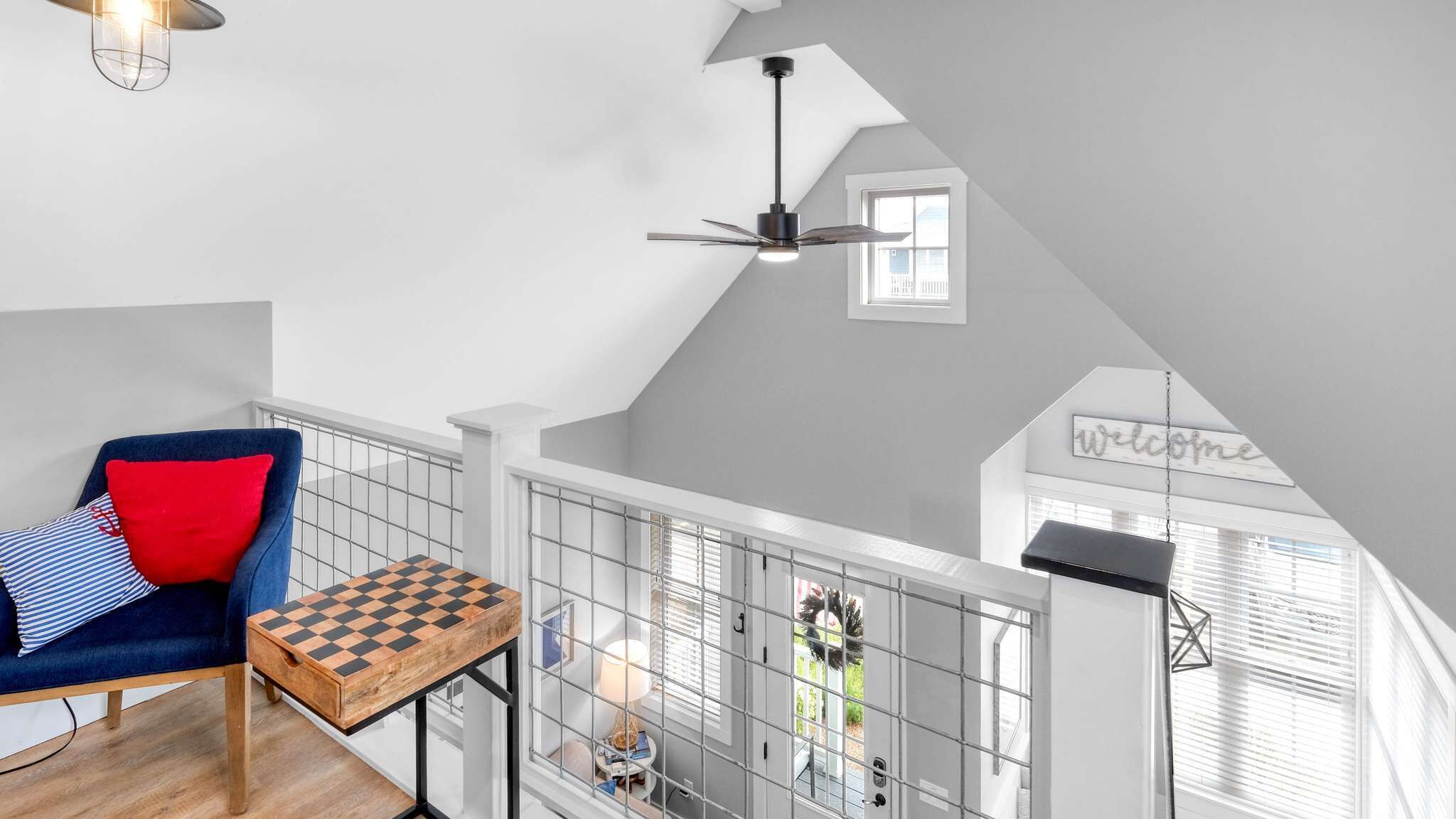 Un espacio tipo loft con techo alto, ventilador de techo, un tablero de ajedrez, una silla azul con cojines rojos y un cartel que dice 'Welcome'.