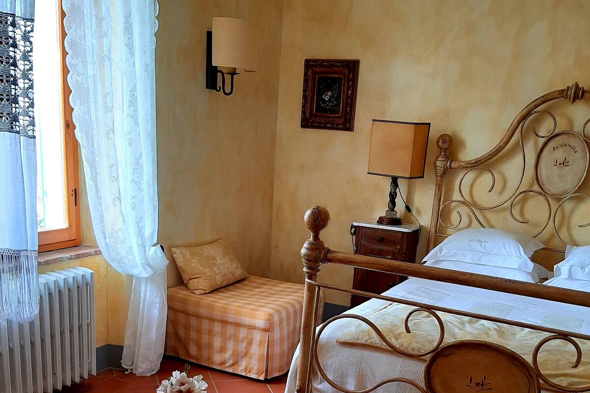 A hotel room with a double bed, a small bedside table with a lamp, a radiator, a window with curtains, and a bedside table with a vase of flowers.