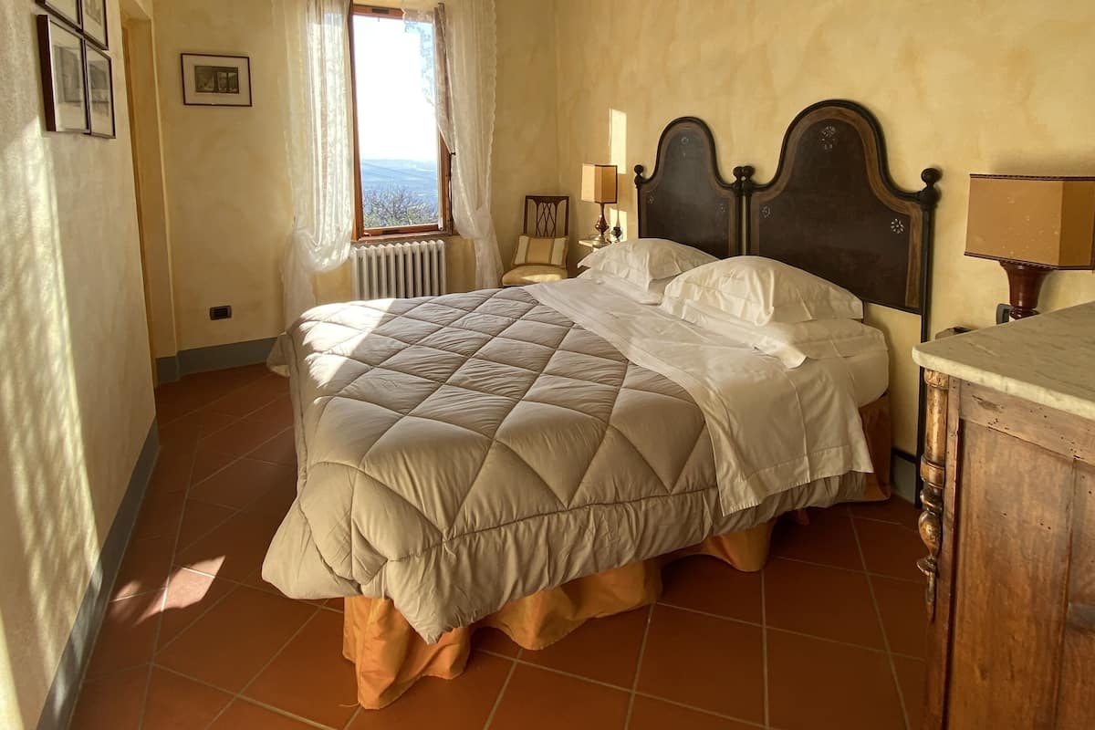 A bedroom with a large bed, a wooden headboard, a nightstand, a lamp, and a view of the outdoors through a window.