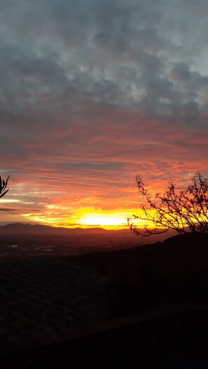 Un atardecer sobre un paisaje montañoso con siluetas de árboles y luces de una ciudad distante.
