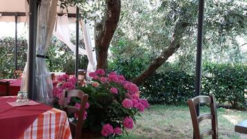Un patio exterior con una mesa y sillas, cubierto por una marquesina, rodeado de follaje y plantas en flor.