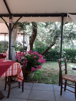 An outdoor patio with a table and chairs, covered by a canopy, surrounded by greenery and flowering plants. - Borgo La Casetta (Lamporecchio)