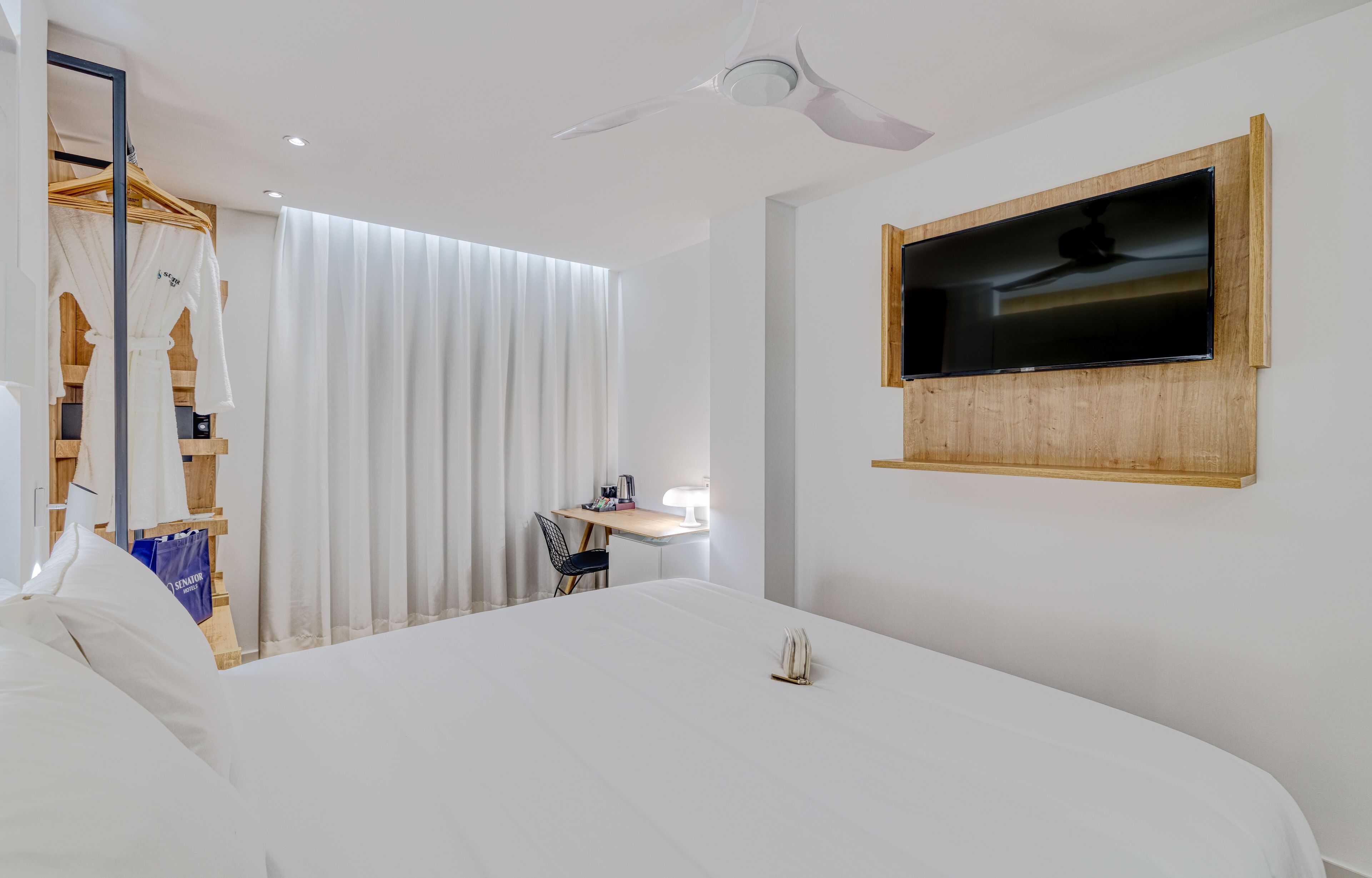 Una habitación de hotel con una cama grande, un televisor montado en la pared, un ventilador de techo y un escritorio con una silla.