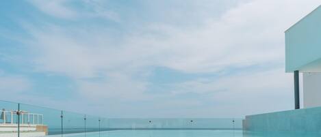 A clear blue sky above a swimming pool with a glass barrier, a white building, and a distant view of the sea.