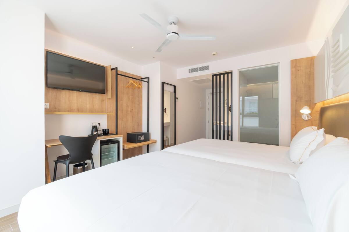 A modern hotel room with a large bed, a desk, a chair, a TV, and a wardrobe.