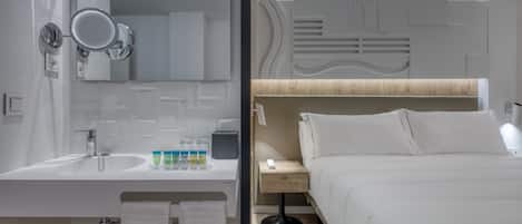 Ein modernes Badezimmer mit Waschbecken, Spiegel und Toilettenartikeln. Angrenzend ein Schlafzimmer mit Bett, Nachttisch und Beistelltisch.
