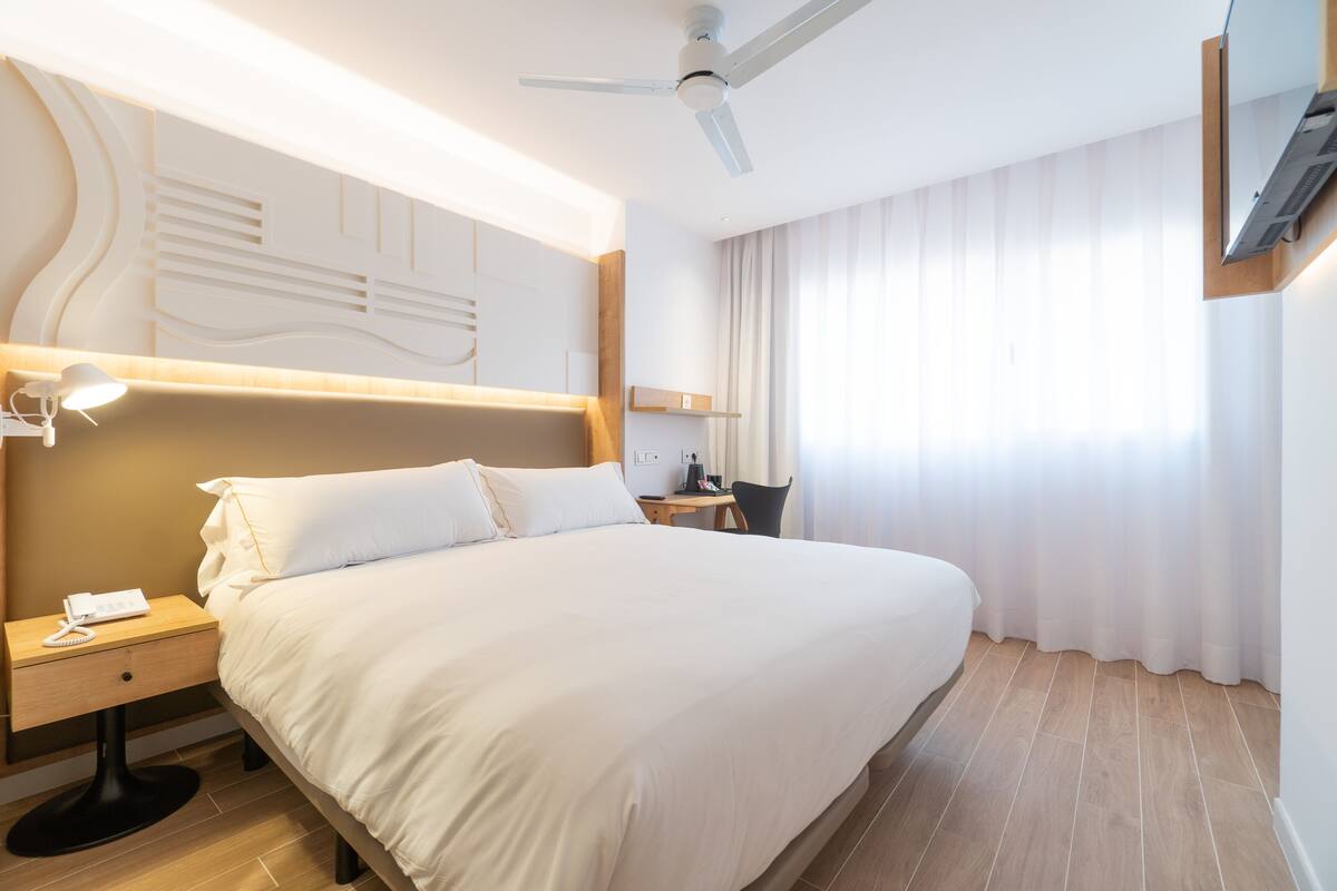 A hotel room with a large bed, a desk, a chair, a desk lamp, a telephone, a ceiling fan, and a TV mounted on the wall.