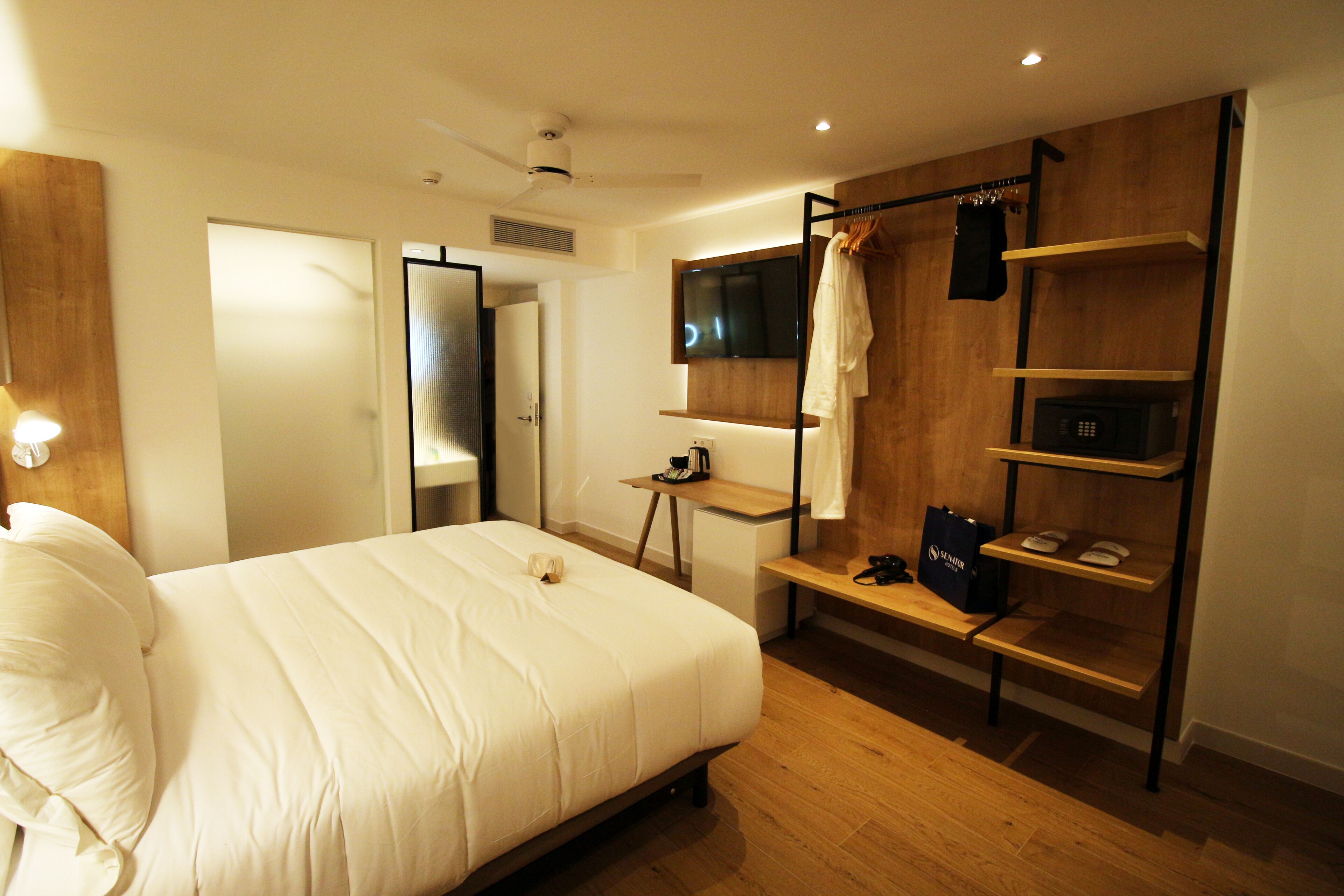 A modern hotel room with a large bed, a desk, a TV, and a wooden shelf with a robe and a bag.