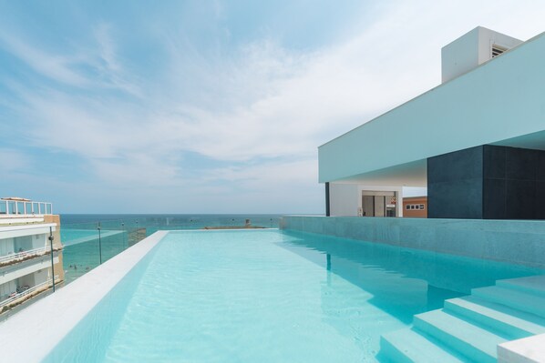 Una piscina infinita moderna con vista despejada al océano, un edificio blanco de techo plano y una escalera que baja a la piscina.