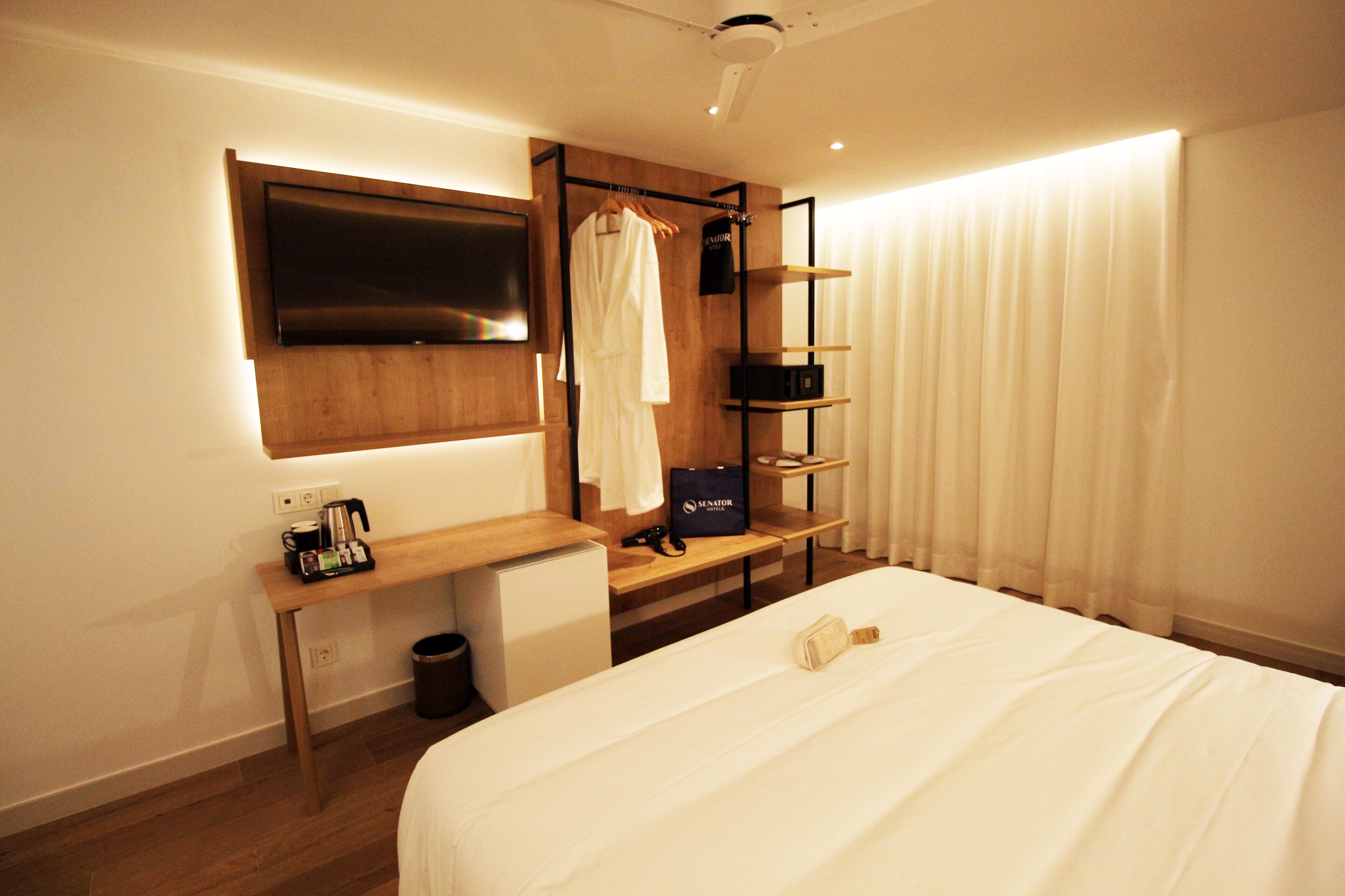 Habitación de hotel con cama, escritorio, televisión montada en la pared, bata colgada en un perchero y ventana con cortinas.