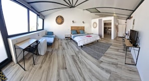 A modern bedroom with a bed, a desk, and a chair. There is a large window and a door leading to another room.