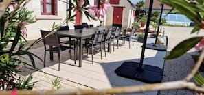 An outdoor patio with a table and chairs, surrounded by potted plants and a building with red shutters.