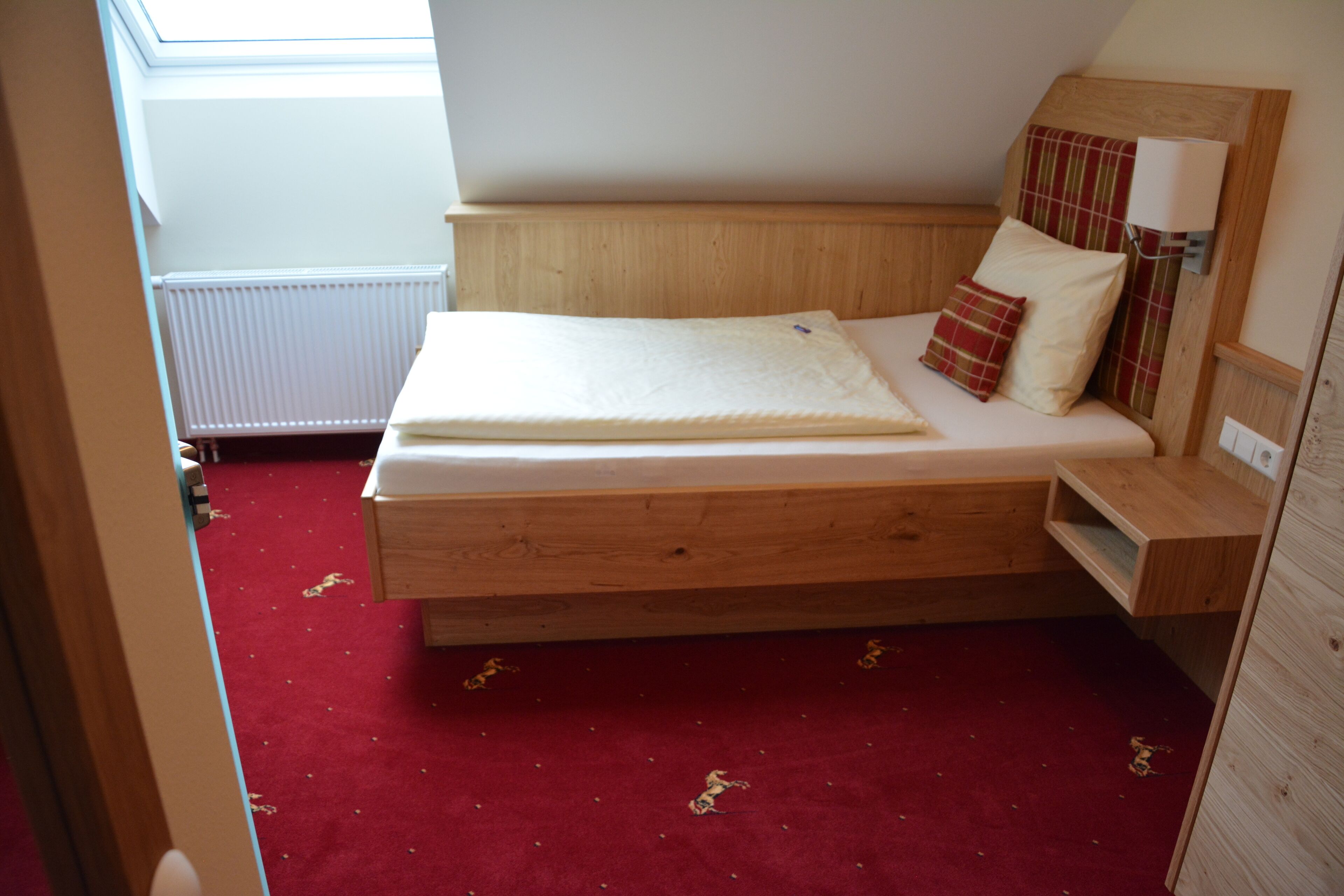 Habitación individual con una cama y cabecera de madera, una mesita de noche y alfombra roja.