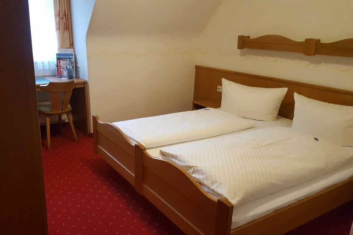 Ein kleines Zimmer mit einem Einzelbett, einem Holz-Kopfteil, einem Schreibtisch mit Stuhl und einem roten Teppichboden.