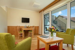 A modern living room with a wooden dining table, green chairs, a flat-screen TV mounted on the wall, and a large window with a view of a staircase and outdoor area.