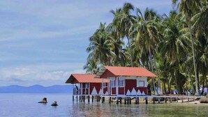 Un edificio de estilo bungalow con techo rojo y paredes blancas, situado sobre pilotes en aguas cristalinas, rodeado de palmeras.