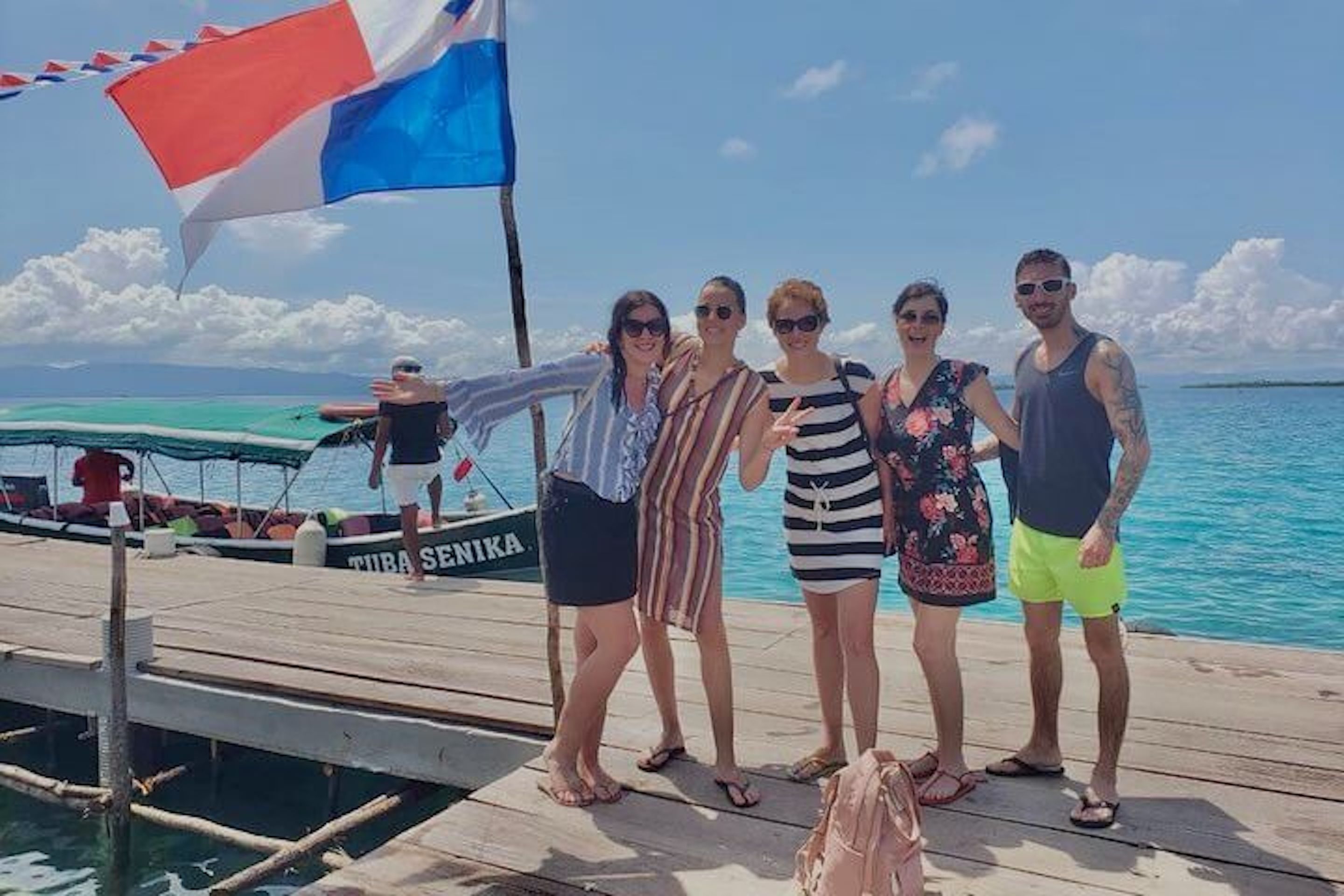 Un groupe de personnes se trouve sur une jetée en bois, au bord de l’eau, et on aperçoit au loin un bateau portant le nom « THIRA SENIKA ».
