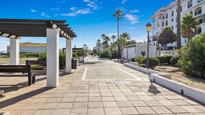 Un sendero pavimentado con bancos, una pérgola y palmeras.