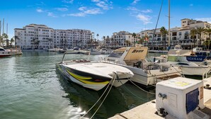 Un puerto deportivo con varias embarcaciones amarradas, un edificio con balcones y palmeras al fondo.