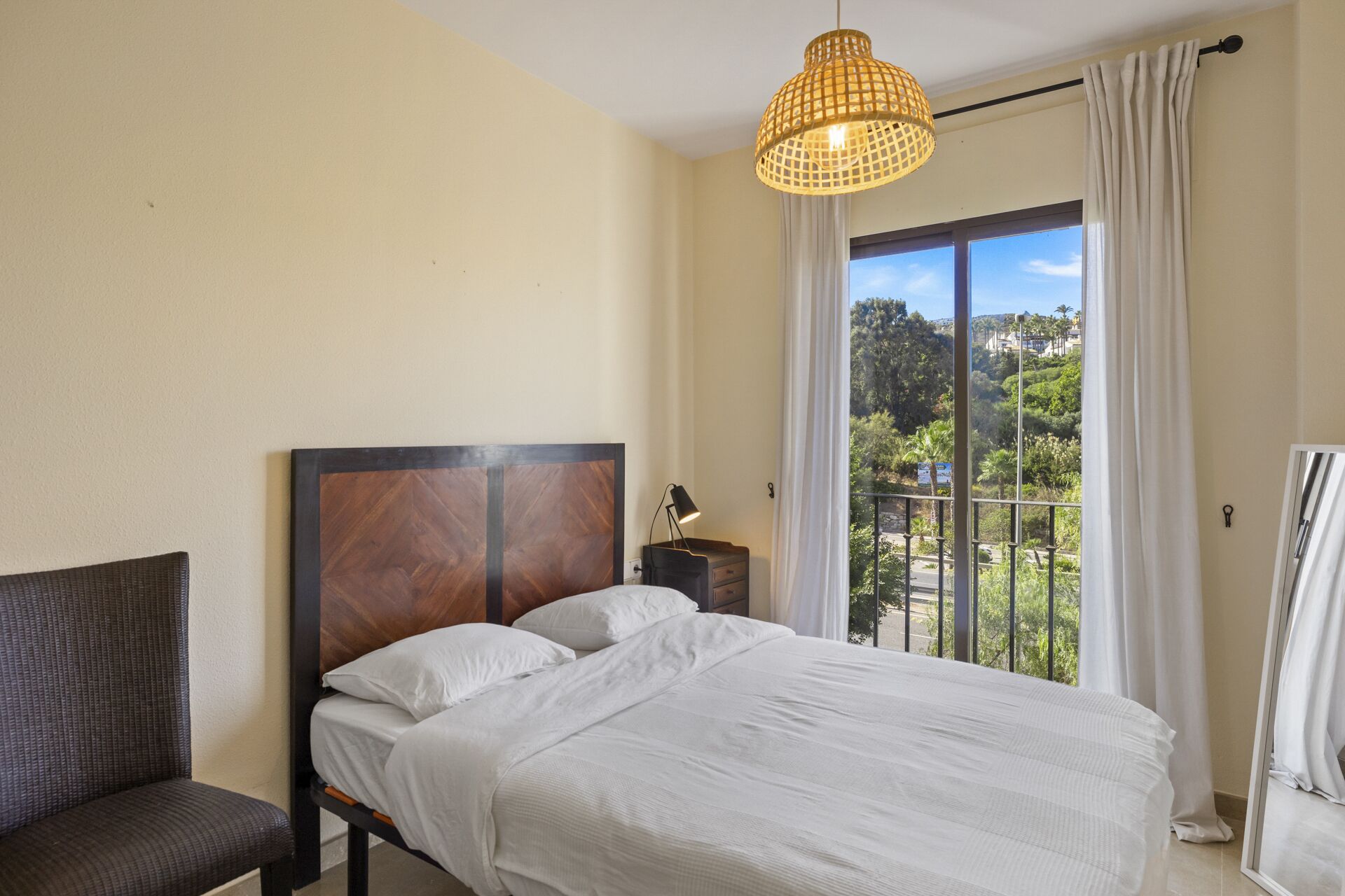 Un dormitorio con cama, una silla, una mesita de noche, una lámpara y vistas al verdor por la ventana.