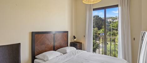 Un dormitorio con cama, una silla, una mesita de noche, una lámpara y vistas al verdor por la ventana.