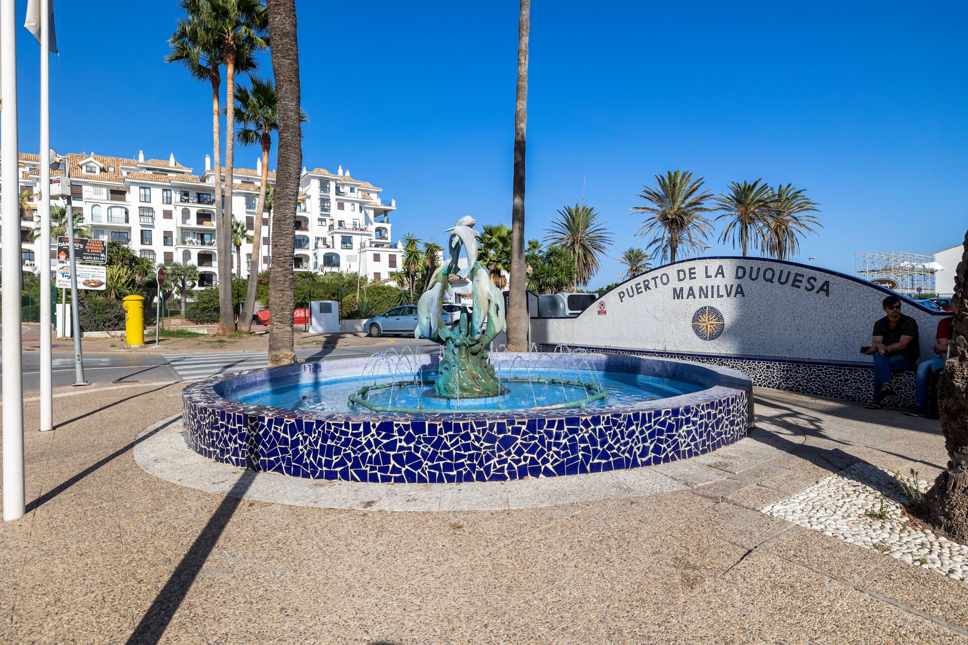 A fountain with a mosaic border, surrounded by palm trees and a sign reading "Puerto de la Duquesa Manila".