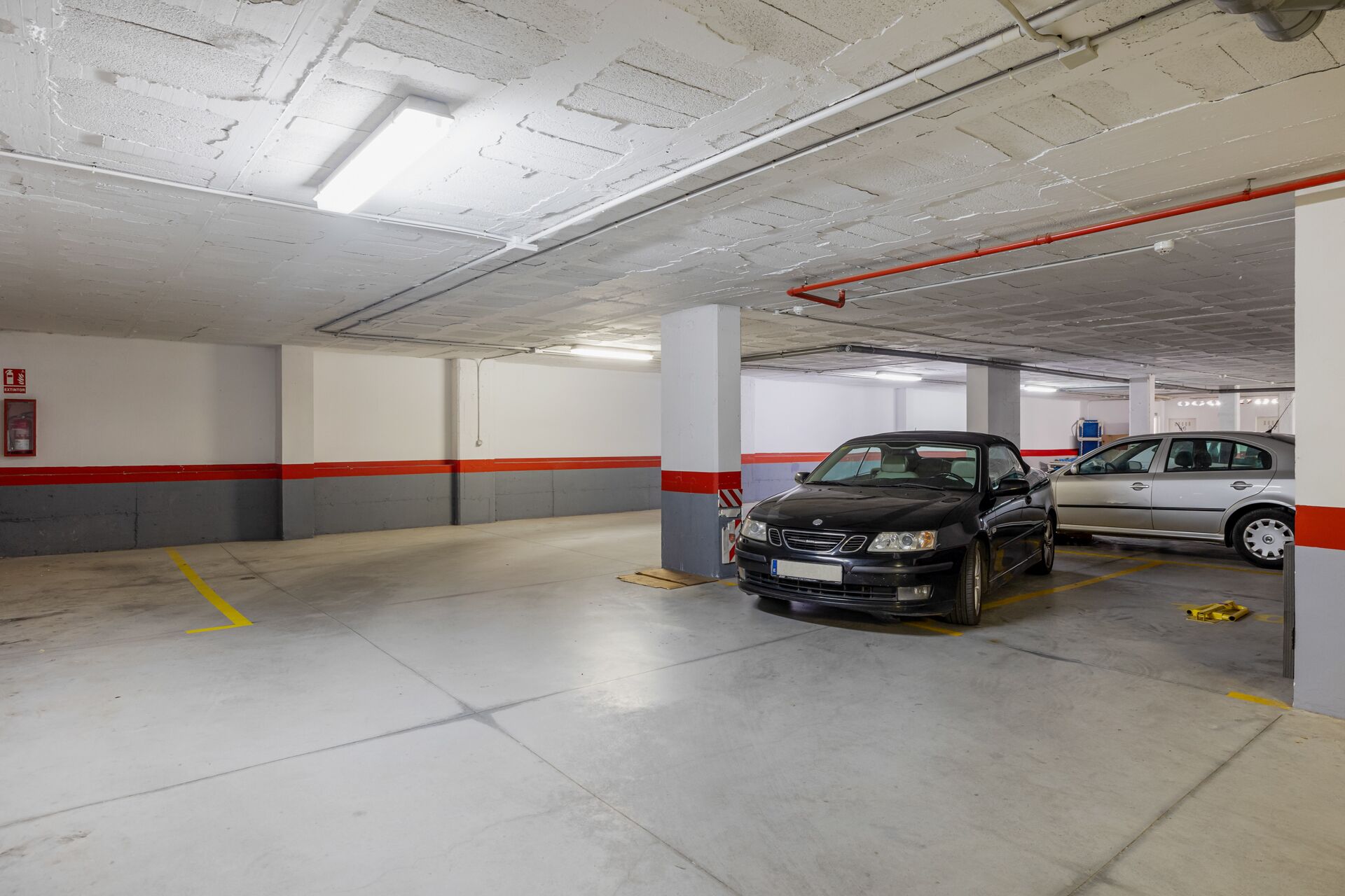A parking garage with two cars parked, concrete floors, and white walls with red stripes.