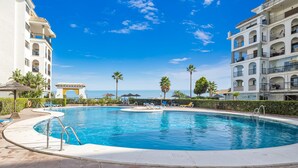 Una piscina con vista despejada al océano, rodeada de palmeras y un edificio blanco con balcones.