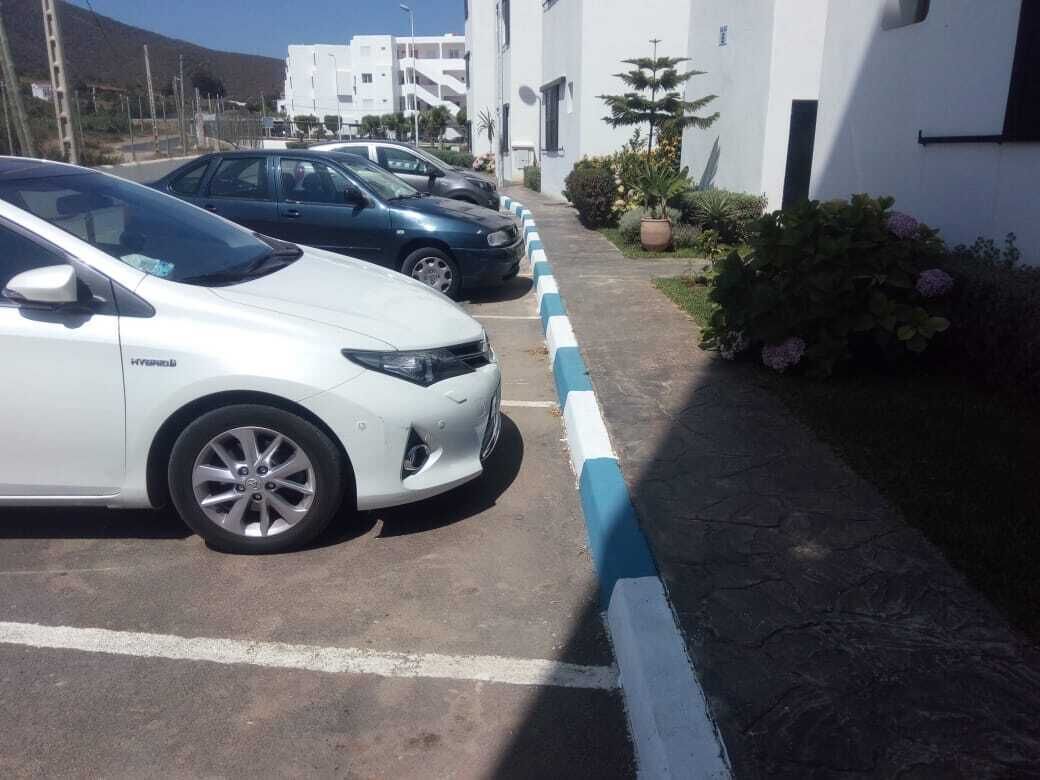 Un espace de stationnement où sont garées plusieurs voitures, dont une voiture blanche sur laquelle on peut lire le mot « HYBRIDE ». En arrière-plan, on aperçoit des bâtiments blancs dotés de balcons et de plantes en pot.