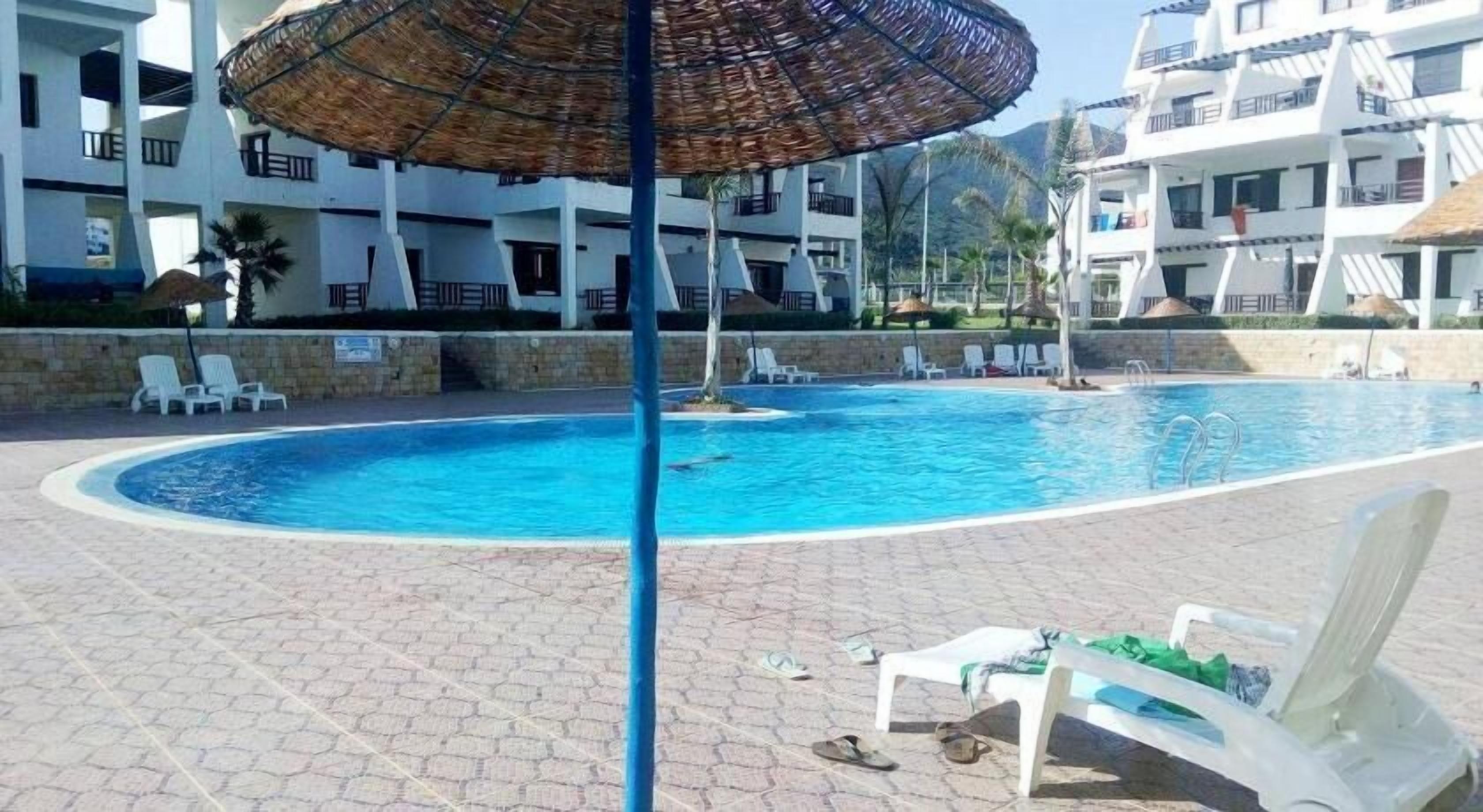 A swimming pool with lounge chairs and umbrellas, surrounded by a stone-paved area and a building with balconies.