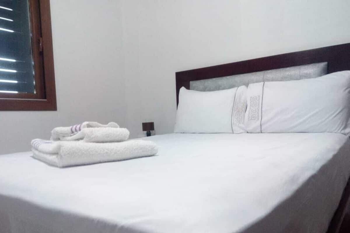 A neatly made bed with white linens and pillows, a wooden headboard, and a window with blinds.