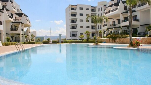 Una piscina rodeada de edificios de apartamentos modernos con balcones, cielo despejado y algo de vegetación.