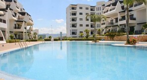 A swimming pool surrounded by modern apartment buildings with balconies, a clear sky, and some greenery. - Résidence Tamuda Golf (M'diq)