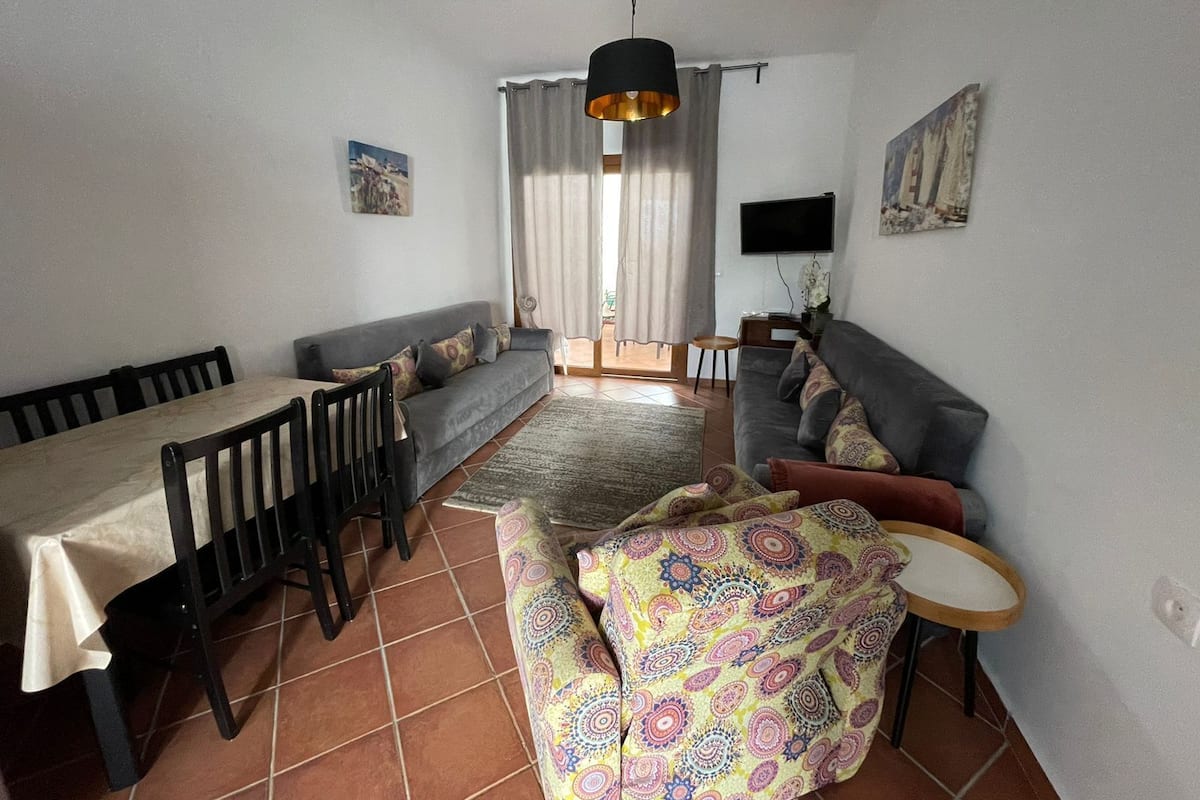 A living room with a sofa, a dining table, chairs, a patterned ottoman, a TV, and a window with curtains.