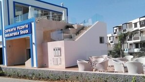 Un edificio moderno con fachada azul y blanca, una escalera que conduce a un balcón y mobiliario exterior con sillas blancas.