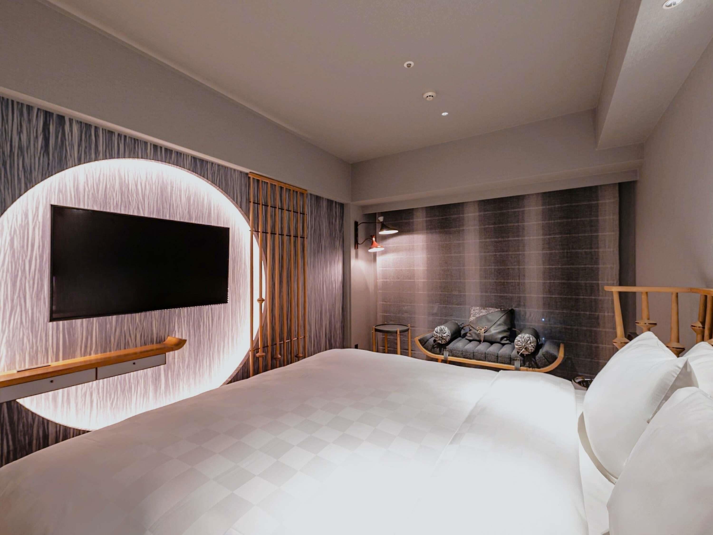 Une chambre moderne avec un grand lit, un téléviseur fixé sur un mur circulaire, un coin salon avec un fauteuil et une petite table, et une lampe fixée au mur.