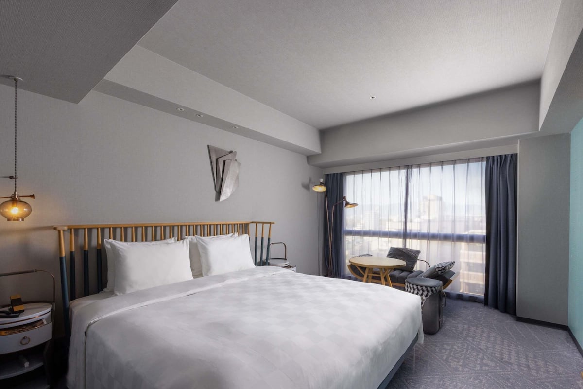 Una habitación de hotel con una cama grande, una mesita pequeña, una silla, un escritorio y un ventanal con cortinas.