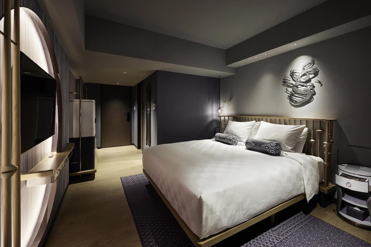 Una habitación de hotel moderna con una cama grande, un televisor, una mesita de noche y una lámpara fijada a la pared.
