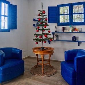 Une pièce avec une table en bois, deux chaises bleues, un sapin de Noël décoré de boules rouges et des volets bleus aux fenêtres.