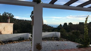 Ett takförsett utomhusområde med en vit pergola, grusunderlag och utsikt över ett kuperat landskap med träd och en sjö eller liknande vattendrag i fjärran.
