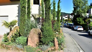 Une rue résidentielle bordée de maisons, dont l’une se distingue par son toit recouvert de tuiles rouges, une fenêtre et un jardin agrémenté de grands arbres et de rochers.