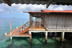 Una cabaña sobre el agua de madera con techo rojo, una terraza de madera y vistas al océano.