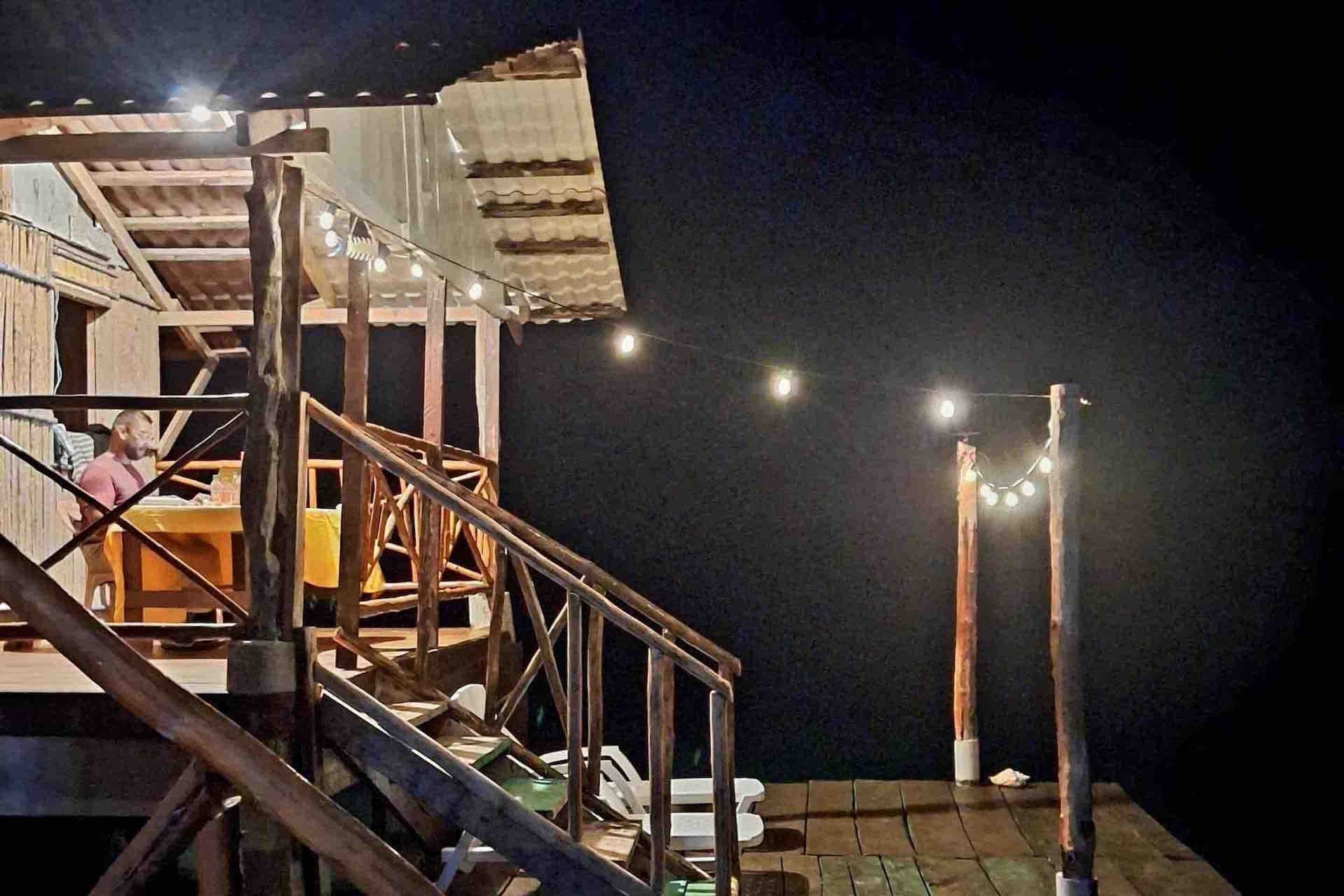 Una escalera de madera que lleva a un balcón con una mesa y sillas, y luces colgantes en la barandilla.