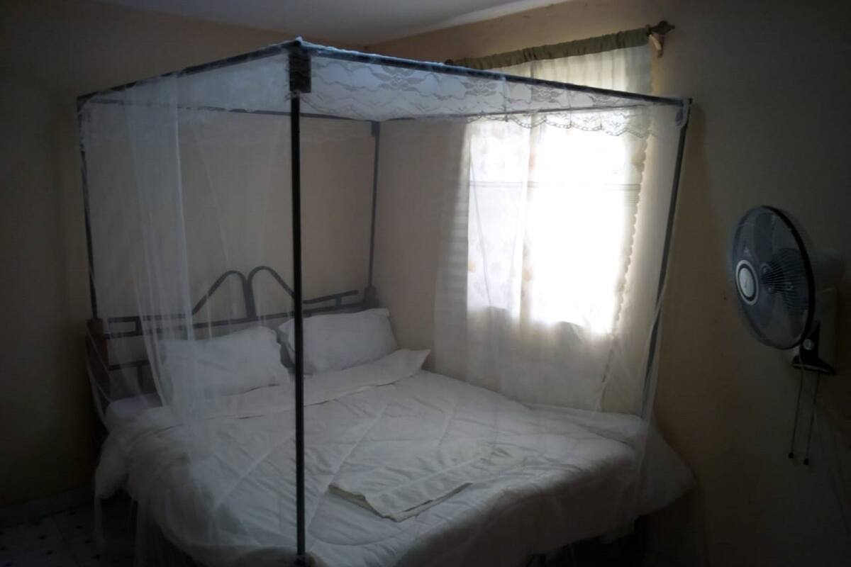 Ein Bett mit Baldachin, ein Ventilator am Nachttisch und ein Fenster mit transparenten Vorhängen.